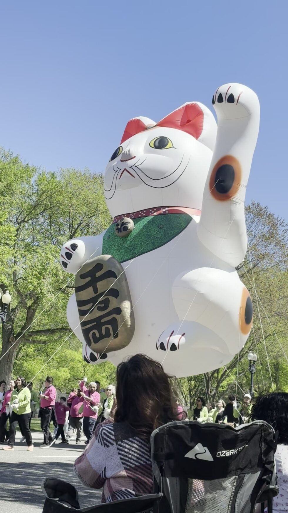 A giant white lucky cat balloon passing spectators in camp chairs along Constitution Avenue