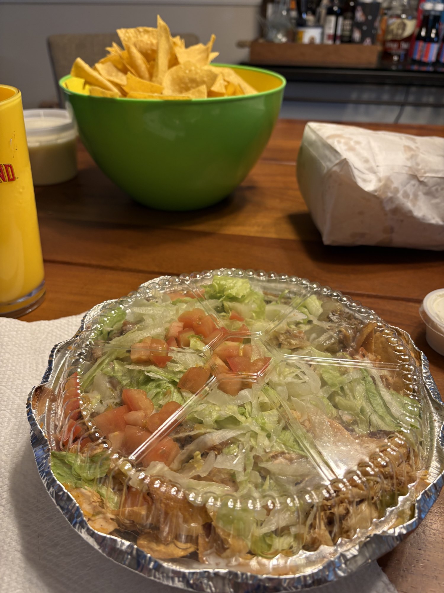A foil tray of Mexican food on a table with a bowl of chips behind it.