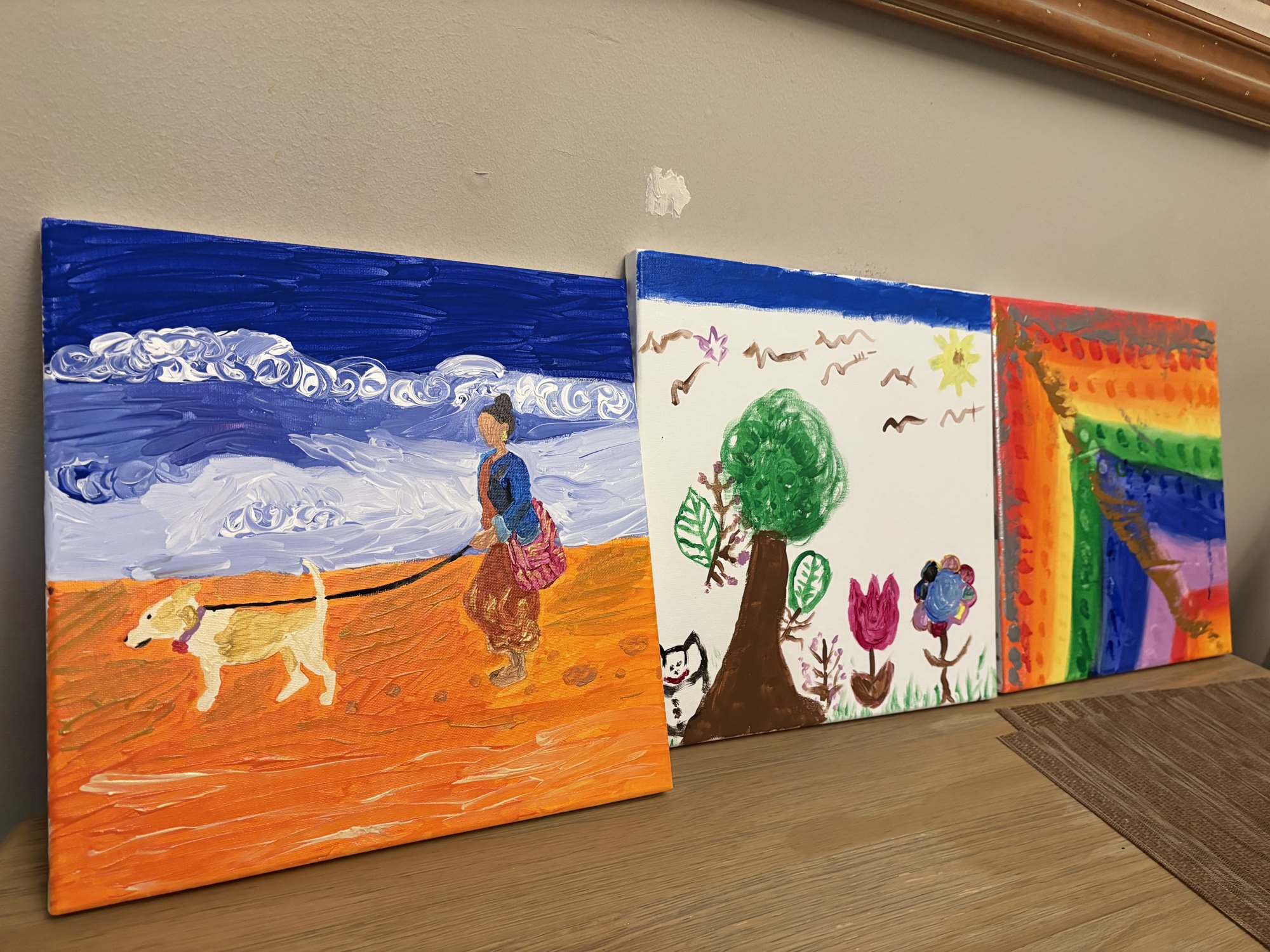 Three painted canvases propped side by side: an orange beach scene, a tree, and a rainbow.