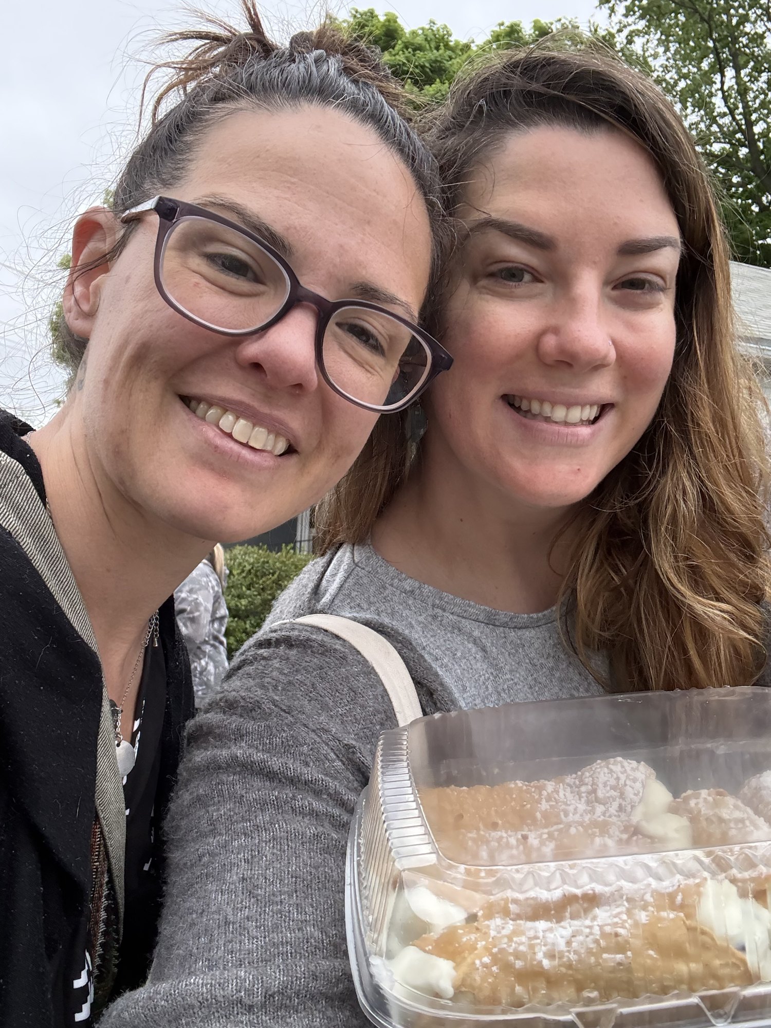 Two friends smiling together, holding a tray of powdered cannoli.