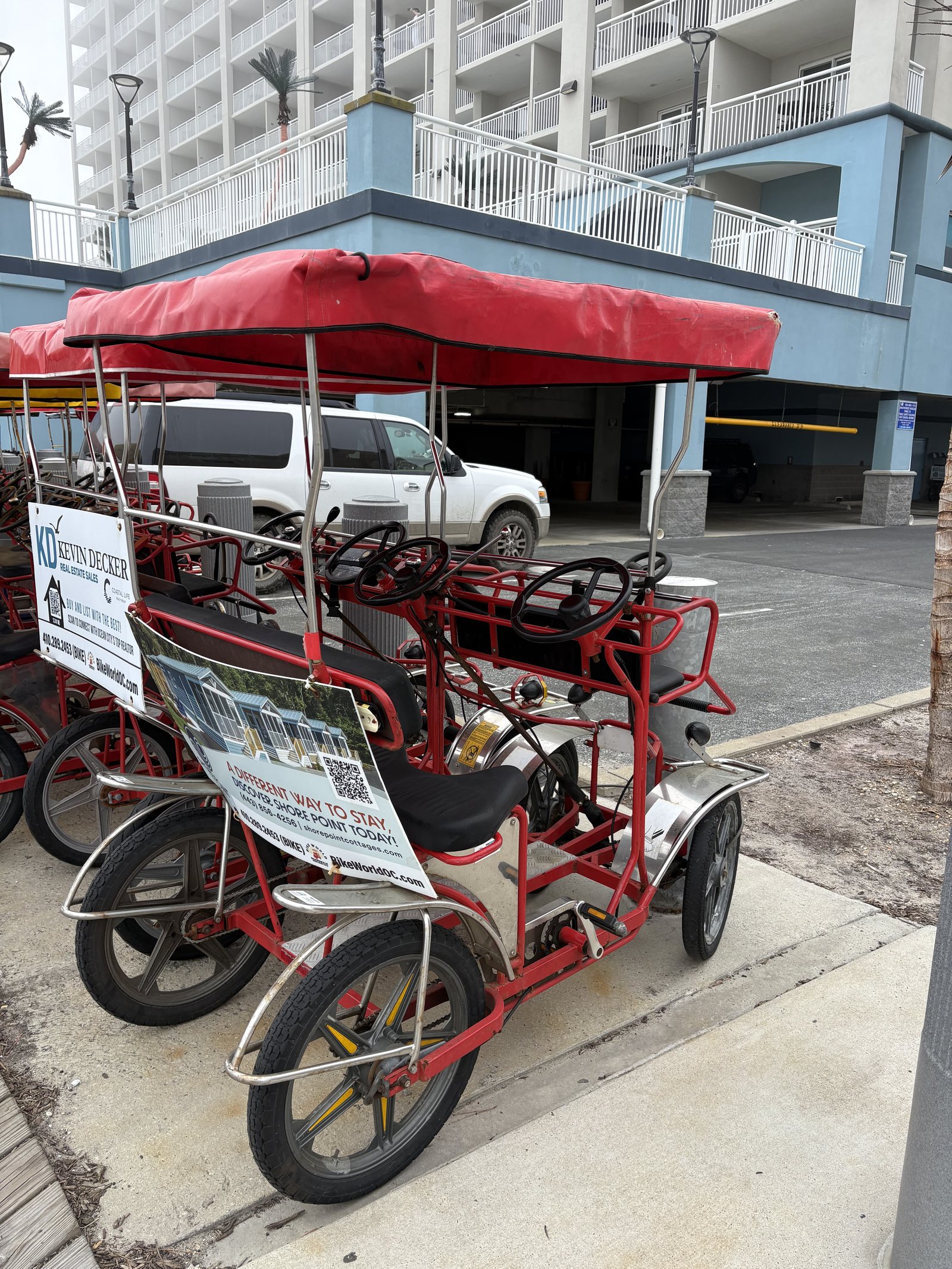 A red four wheeled surrey bike parked on the boardwalk beside a closed hotel