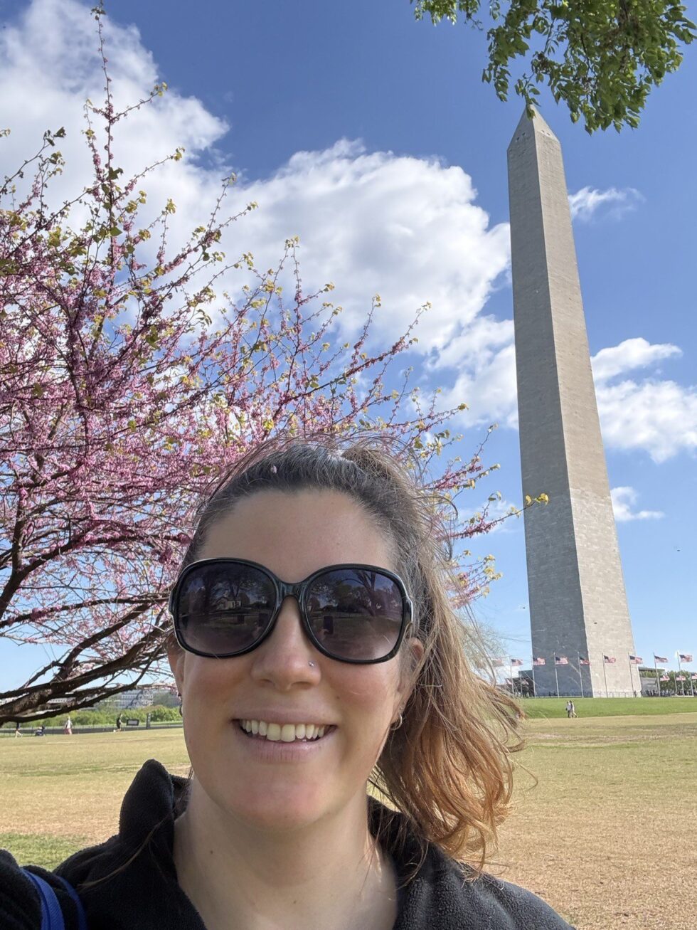 Hannah smiling in sunglasses under a pink flowering tree with the Washington Monument behind her